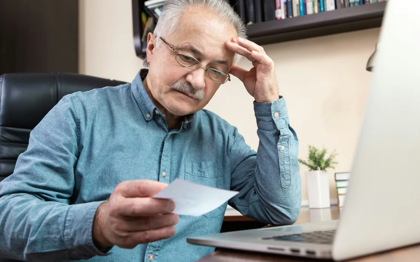 Older man wearing glasses and a denim shirt sitting at a desk, looking thoughtfully at a piece of paper while working on a laptop.