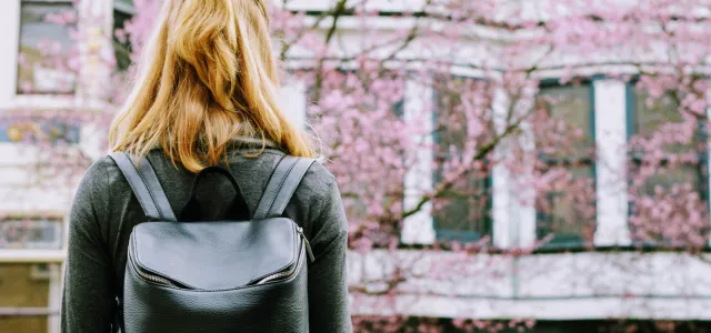 Woman with blonde hair wearing a black backpack, standing outdoors and facing a building with blooming pink trees in the background.