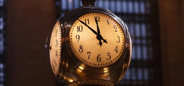 Ornate brass clock showing the time as 11:55, set against a background of tall windows with metal grilles, likely inside a historic train station or building.