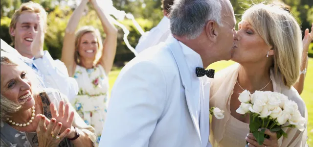 Older couple sharing a kiss at their wedding while guests clap and celebrate outdoors on a sunny day.