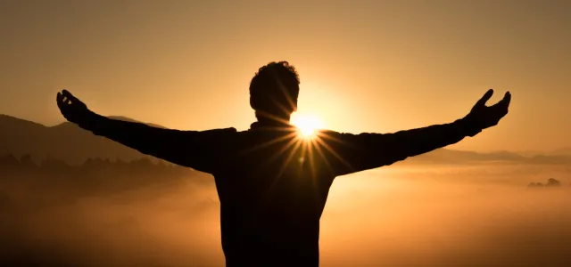 Person standing with arms outstretched facing the sunrise, silhouetted against a golden sky and misty landscape, symbolizing freedom or gratitude.