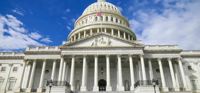 Front view of the United States Capitol building with its grand staircase, columns, and white dome under a partly cloudy blue sky.
