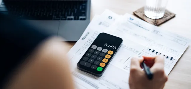 Person reviewing financial documents at a desk with a smartphone calculator, laptop, and glass of water, symbolizing budgeting or expense management.