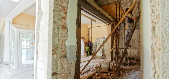 Interior of a building under renovation with exposed brick walls, debris on the floor, and wooden supports holding up partially demolished sections.
