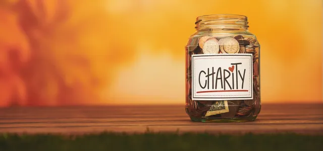 Glass jar labeled “Charity” filled with coins and cash, placed on a wooden surface with a warm orange background symbolizing generosity and giving.