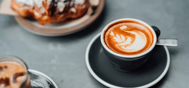 Cup of cappuccino with latte art on a saucer next to a plate with a chocolate croissant on a gray table.