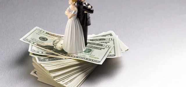 Bride and groom wedding cake toppers standing on a stack of U.S. dollar bills, symbolizing the financial aspects of marriage.