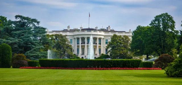 The White House in Washington, D.C., viewed from the lawn with trees and flowers in the foreground under a partly cloudy sky.