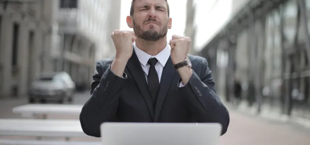 Man in a suit sitting outdoors at a table with a laptop, raising his fists and appearing excited or triumphant.