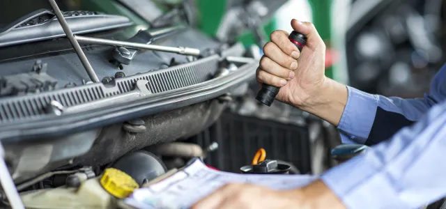 A person looking under the hood of a car. They have a flashlight and a checklist. West Financial Services.